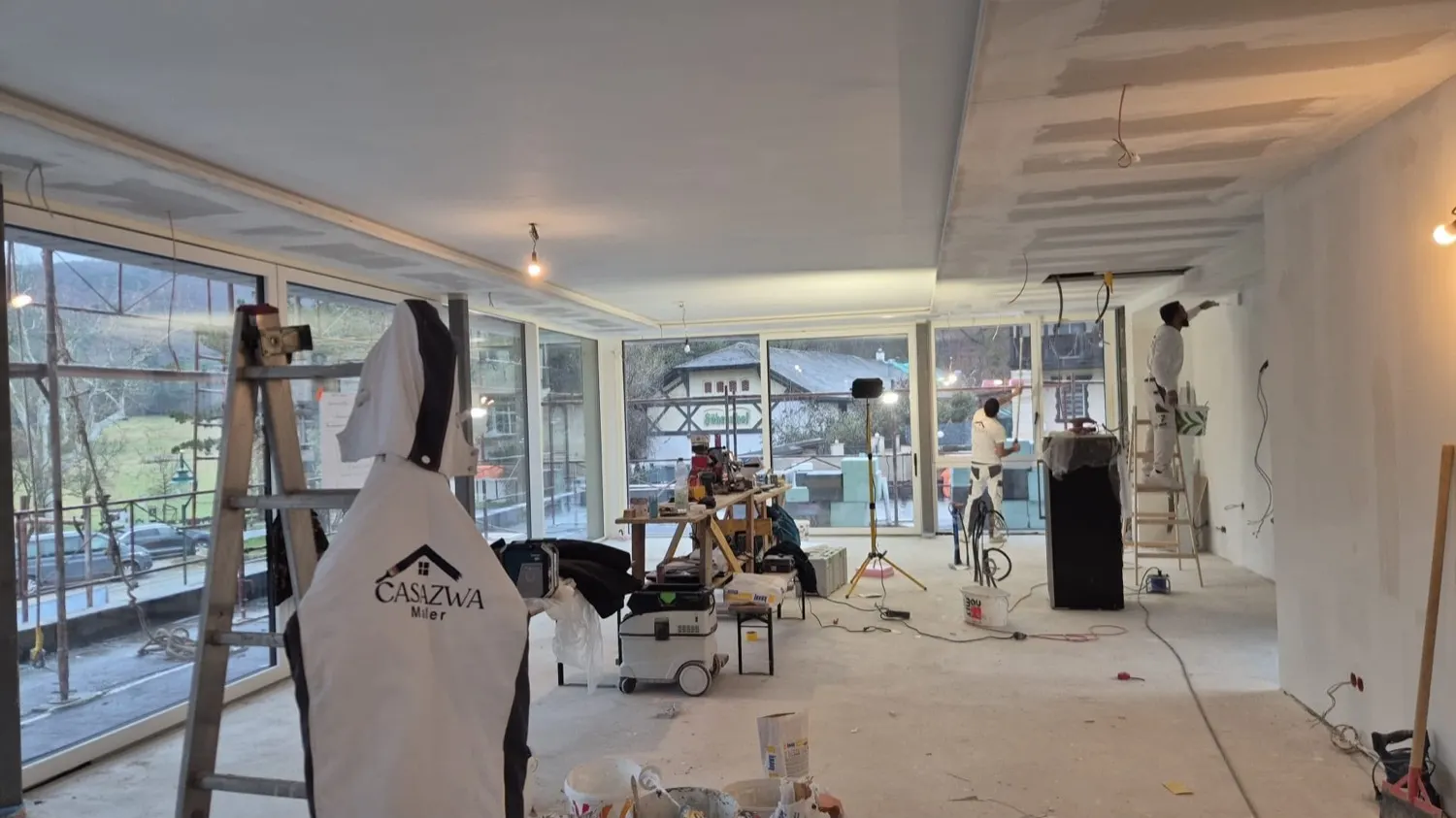 Baustelle von Casa Zwa: Arbeitsjacke mit Firmenlogo an der Leiter, dahinter frisch gespachtelte Wände und die Fensterfront